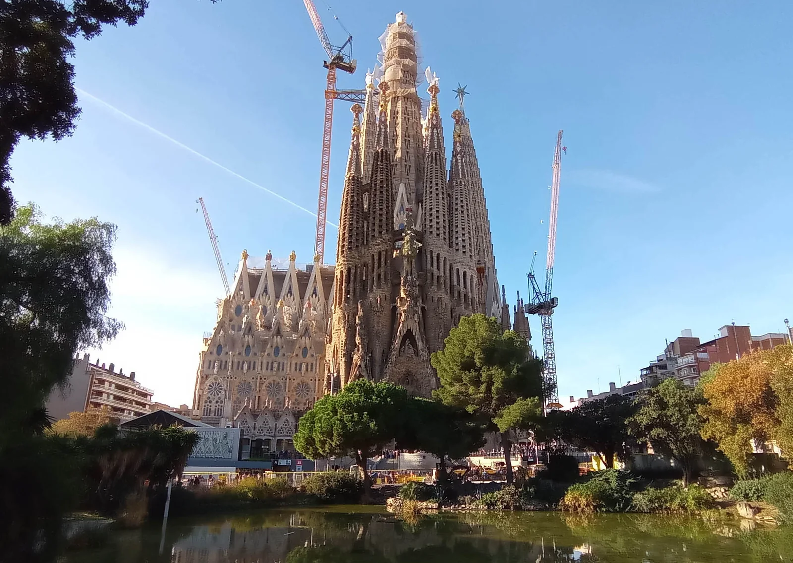 Sagrada Família’s eastern façade and towers above the trees and pond of Plaça de Gaudí, with construction cranes
