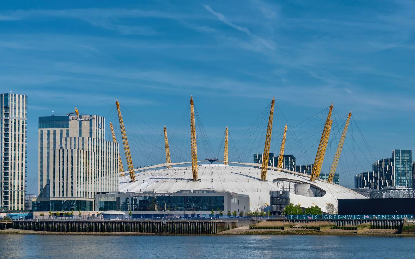 The white canopy and yellow masts of The O2 beside the Thames, with an adjacent riverside hotel